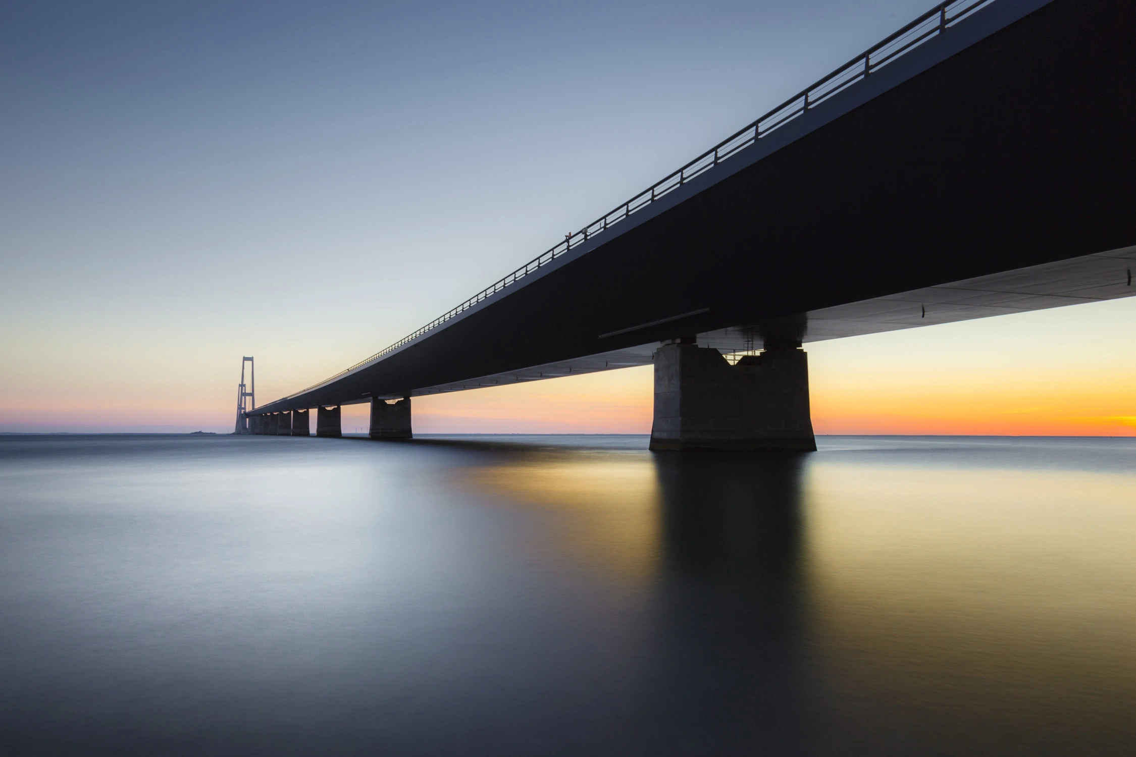 Outdoor image of a bridge suspended over a body of water, with the sun setting in the background to the right side of the image.