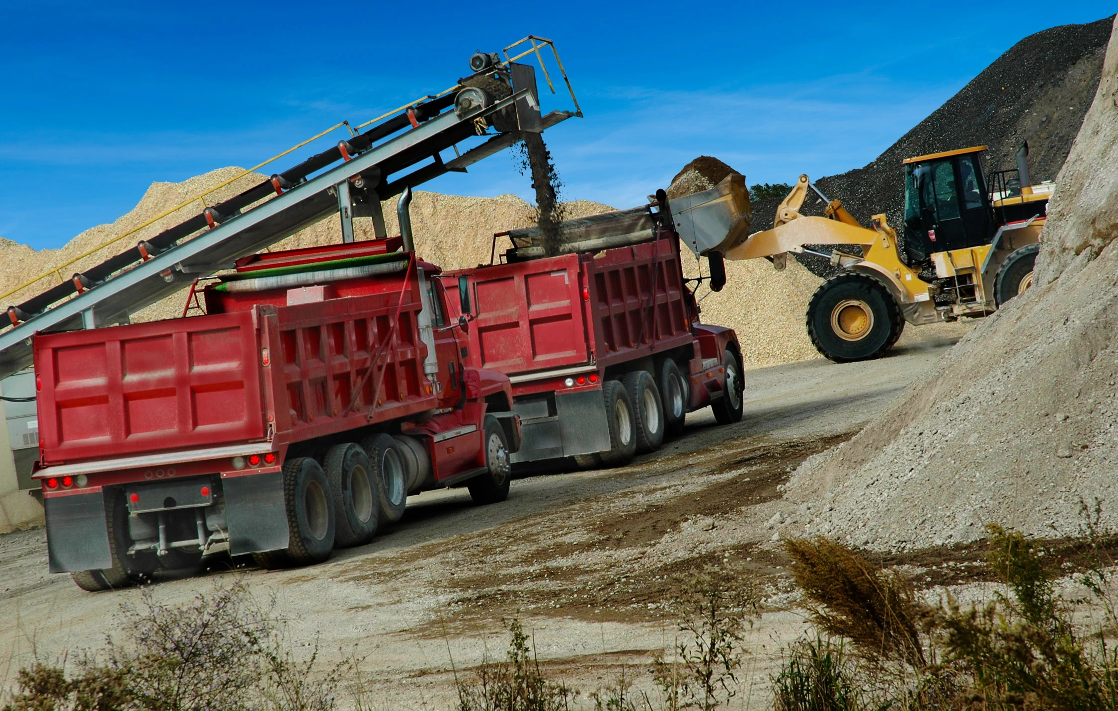 Image of two red dump trucks at a construction site receiving material from a conveyor with some earth-moving equipment in the background.